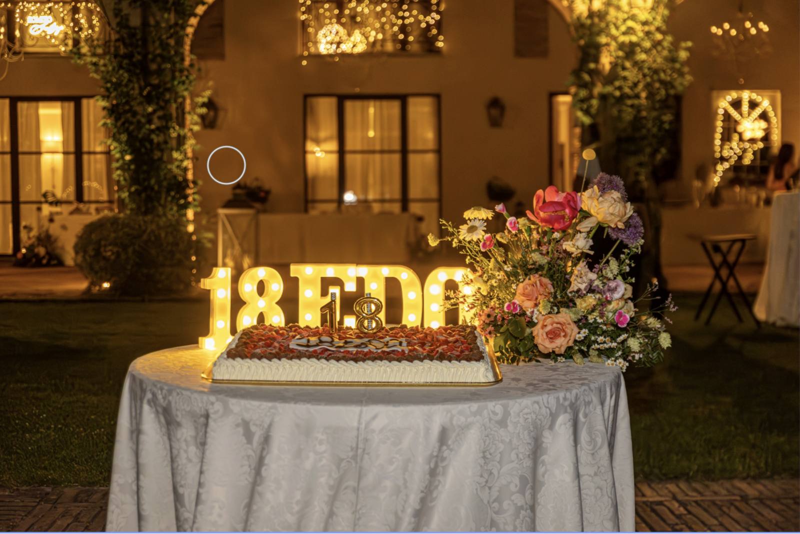 Tavolo con torta di compleanno, lettere luminose '18' e composizione floreale colorata in giardino di sera