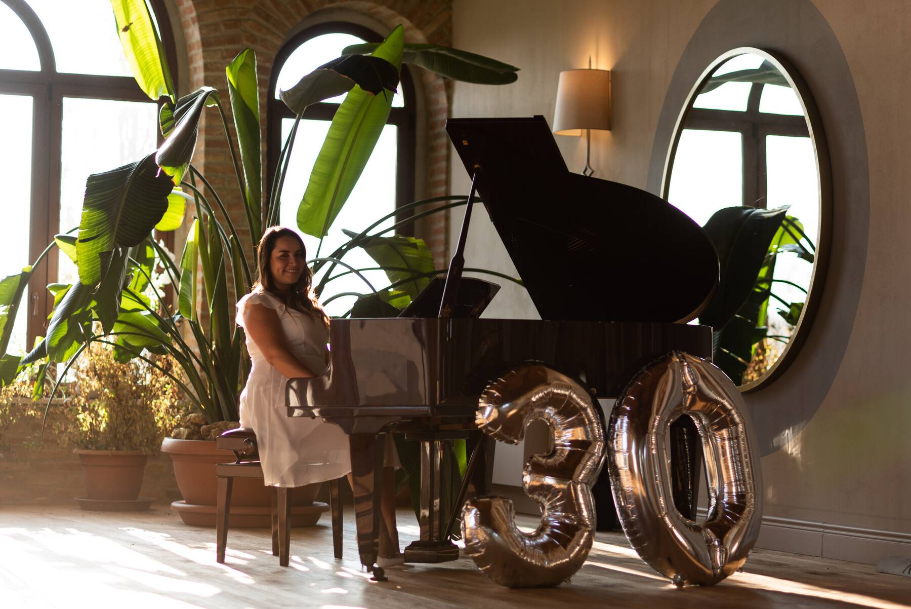 Festeggiata al pianoforte a coda con palloncini dorati '30' per il compleanno, tra le piante e il finestrone ad arco del Salone del Camino