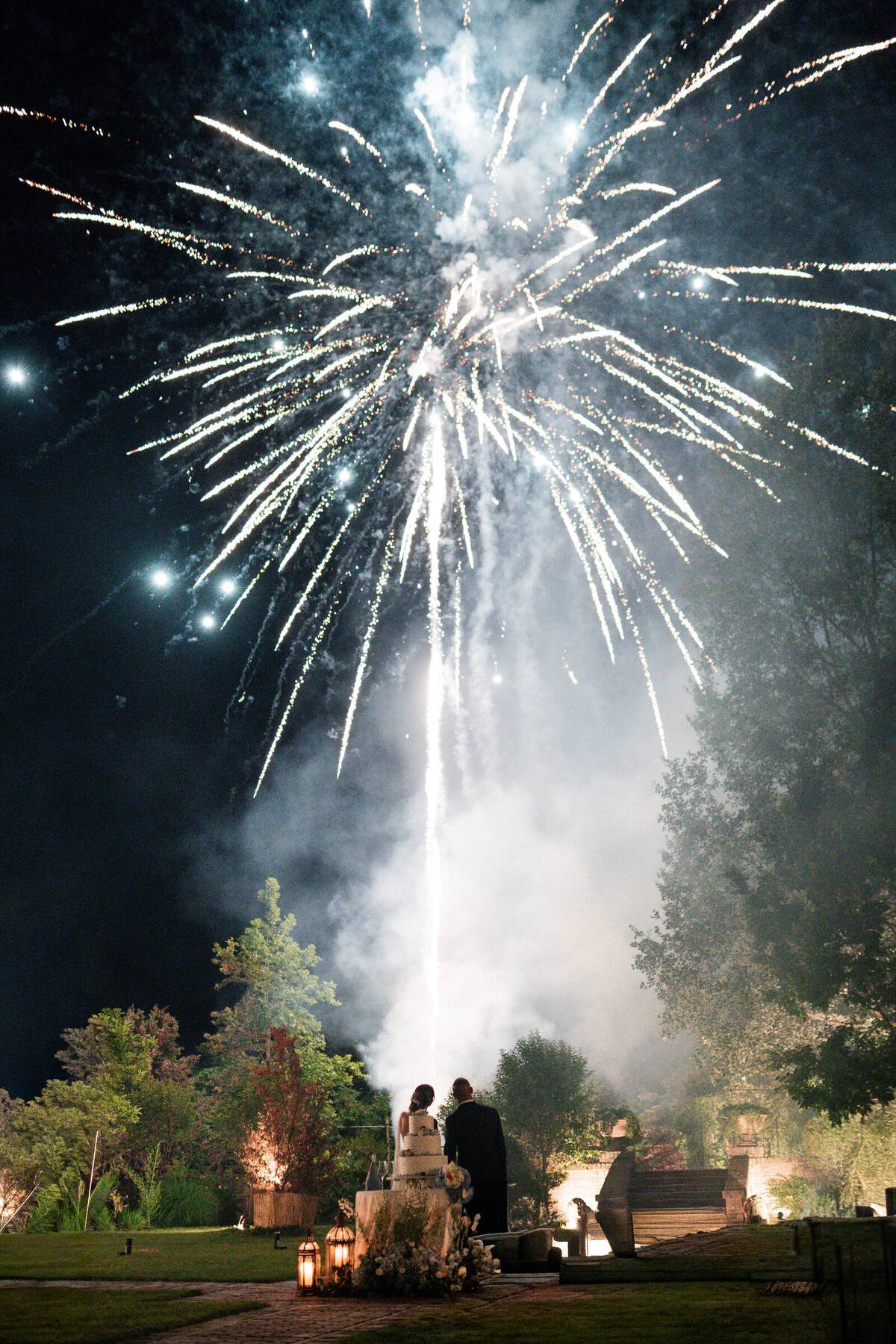 Gli sposi abbracciati davanti alla torta nuziale sotto un cielo di fuochi d'artificio