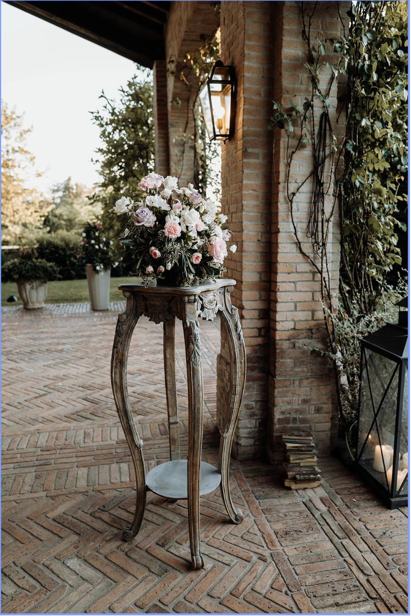 Tavolino antico con composizione floreale rosa e bianca sotto il portico, accanto a una lanterna