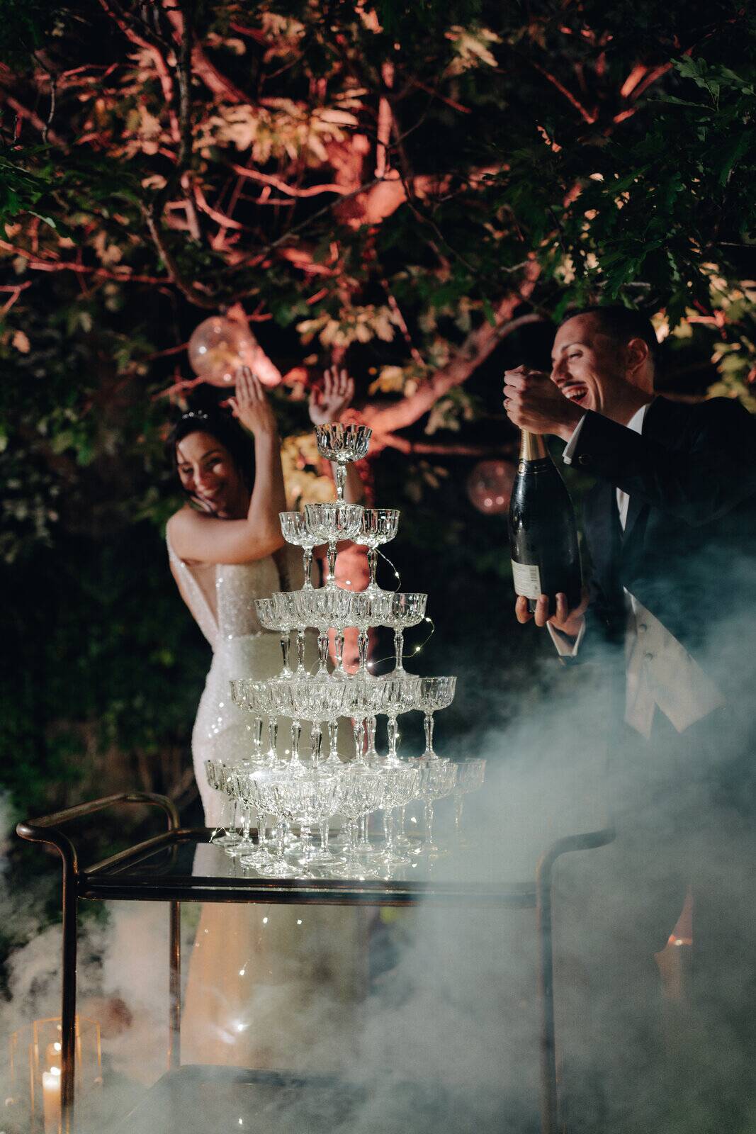 Sposi brindano tra il fumo scenico accanto alla piramide di coppe di champagne