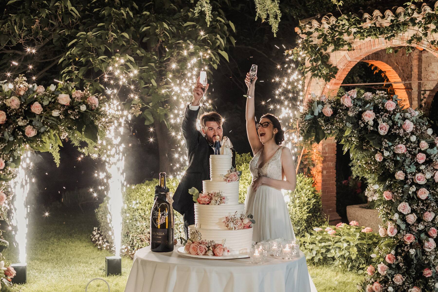 Sposi brindano accanto alla torta nuziale a quattro piani sotto l'arco di rose
