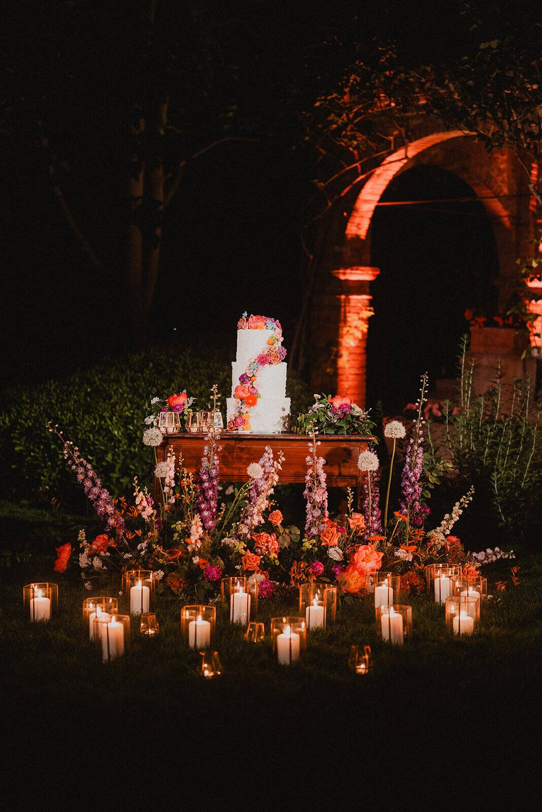 Torta nuziale su tavolo in legno tra fiori colorati e candele, davanti all'arco in mattoni illuminato